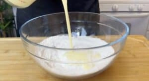 Mixing batter: Pouring liquid into a flour-filled bowl on a wooden surface, baking process in a kitchen setting.